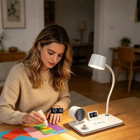 A woman sits at a wooden table in a warmly lit home environment, using a white smart desk lamp with integrated digital clocks and a small heated pad. The lamp is turned on, casting a soft glow, and the woman works on colorful sticky notes on the table.