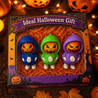 A Halloween-themed gift box containing three small pumpkin-faced character figurines in blue, green, and purple hoodies, displayed on orange crinkle paper inside a decorated box with pumpkins and string lights.