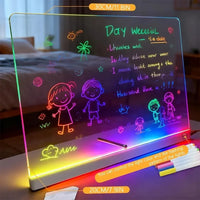 A black LED light-up writing board shown on a bedside table in a bedroom setting, with colorful neon-style drawings and text on the black surface. The board has a glowing multicolor LED light border and a pen resting on it, with promotional text overlays indicating dimensions and features.