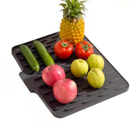 A black rubber kitchen draining mat with a slotted, ribbed surface, shown on a white background with various fruits and vegetables placed on top, including a pineapple, cucumbers, tomatoes, apples, and pears.
