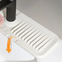A white plastic sink drain guard with slotted ridges, shown installed under a faucet with water flowing into the sink. A hand is visible on the left holding the faucet, and an orange arrow graphic illustrates water diversion.