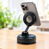 A black magnetic phone stand with a suction cup base, holding a silver smartphone on a wooden table with a blurred indoor background.